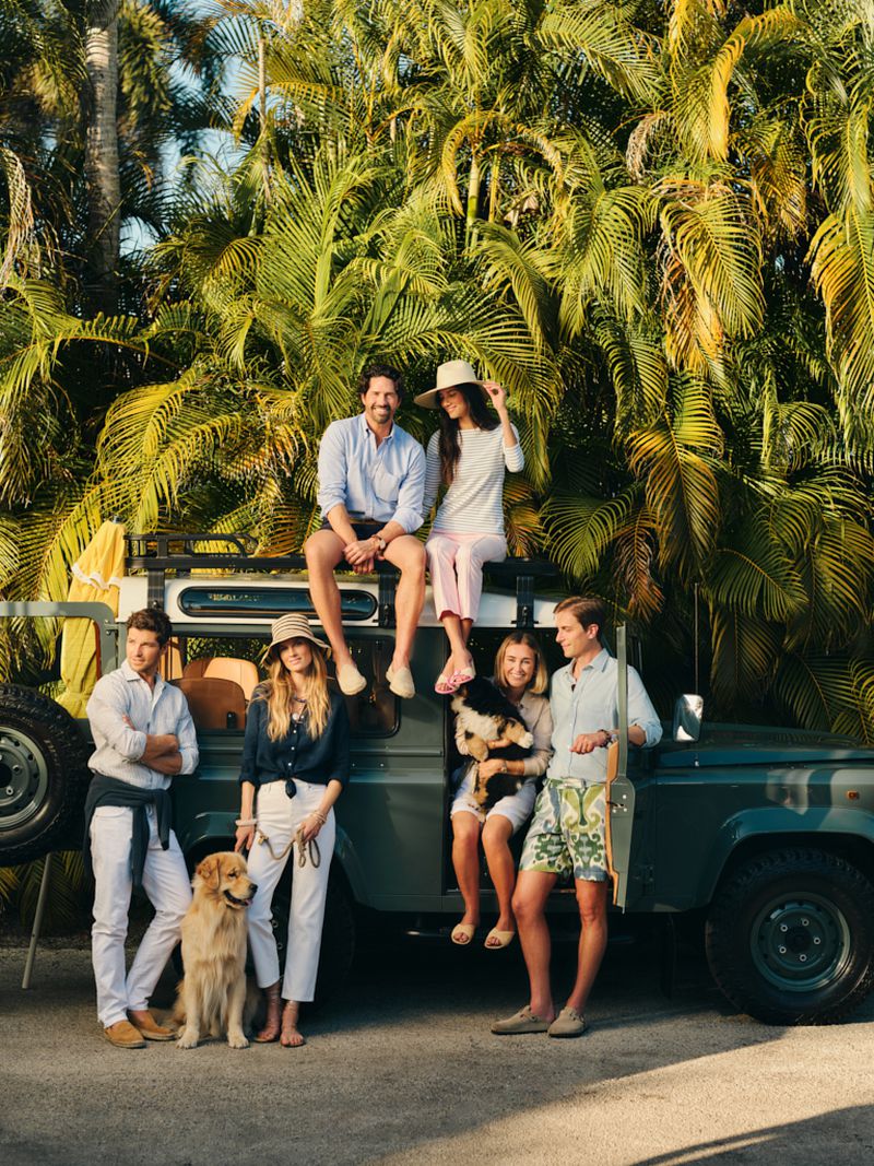 Six models and two dogs pose around a vintage dark green 4x4 vehicle against a lush backdrop of tall palm trees in warm sunlight.