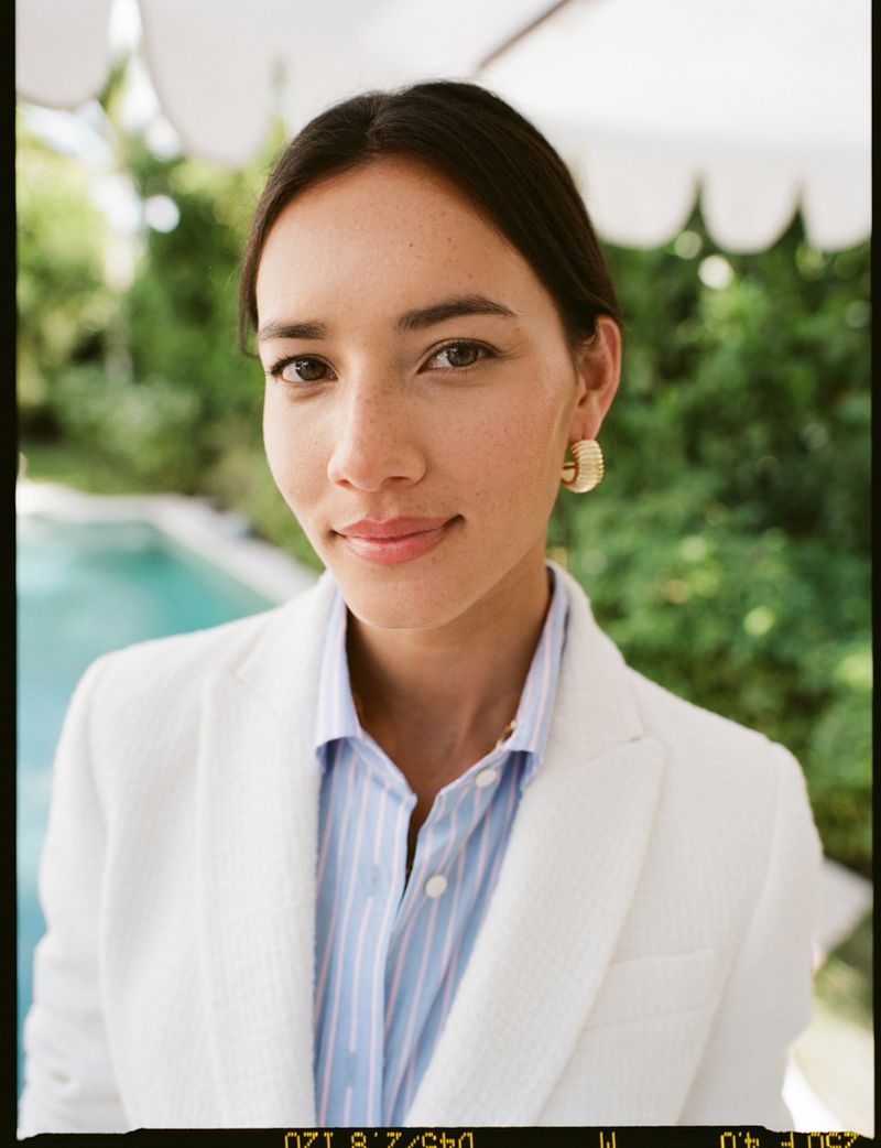 Close-up portrait of a woman in a white blazer and light blue striped shirt wearing gold earrings outdoors by a swimming pool.