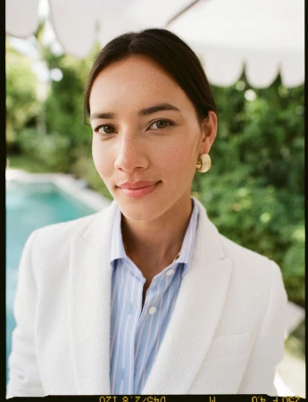 Close-up portrait of a woman in a white blazer and light blue striped shirt wearing gold earrings outdoors by a swimming pool.