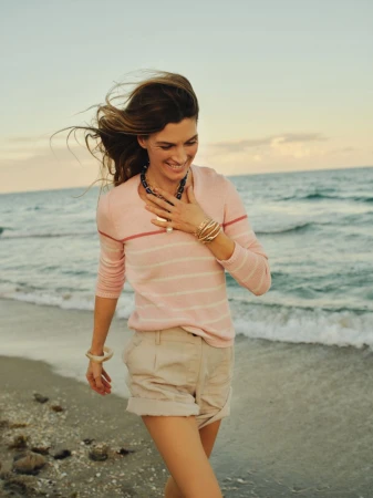 Smiling woman walking along a sandy shoreline wearing a pink striped sweater and beige shorts.