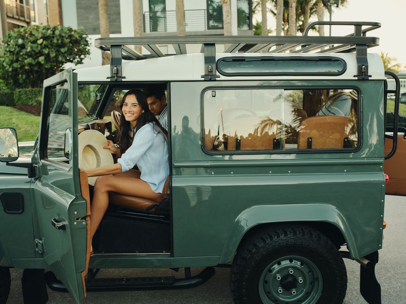 Smiling woman seated inside a vintage green off-road vehicle holding a wide-brim sun hat with a man seated beside her.