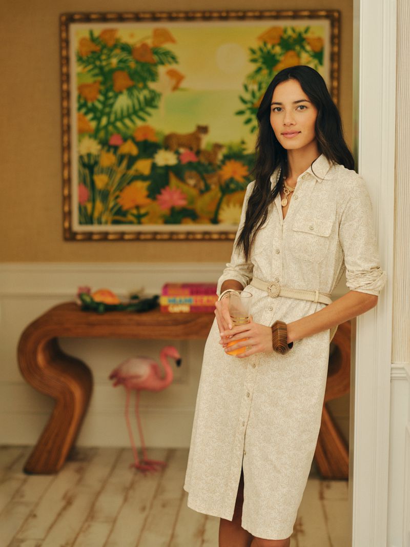 A woman in a cream collared belted shirt dress stands leaning against a doorway while holding a drink in an interior room.