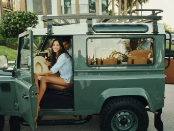 Smiling woman seated inside a vintage green off-road vehicle holding a wide-brim sun hat with a man seated beside her.