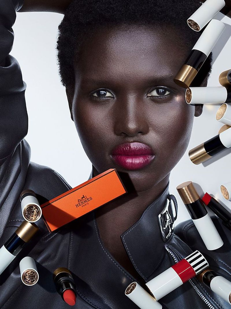 Amar Akway wearing bold berry lipstick and a black leather jacket surrounded by floating Rouge Hermès lipsticks against a white background.