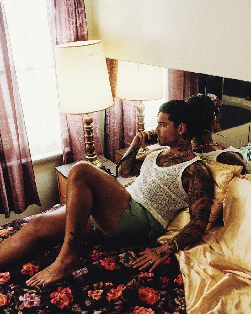 Maluma reclining on a bed holding a vintage telephone receiver in a hotel room for Vogue Hombre.