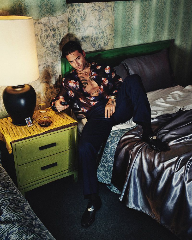Maluma seated on a bed holding a remote control in a retro bedroom styled with vintage wallpaper and a bedside lamp.