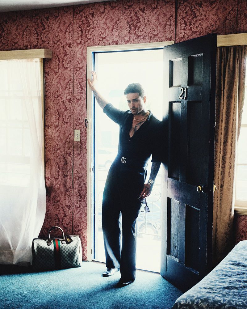 Maluma posing in a vintage hotel room doorway wearing an all-black outfit and Gucci belt with luggage resting nearby on the floor.