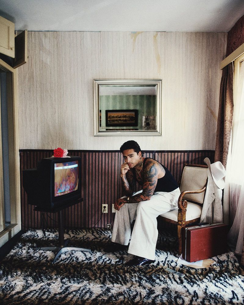 Maluma wearing a black tank top and wide white trousers seated in an armchair beside a vintage television in a retro room.