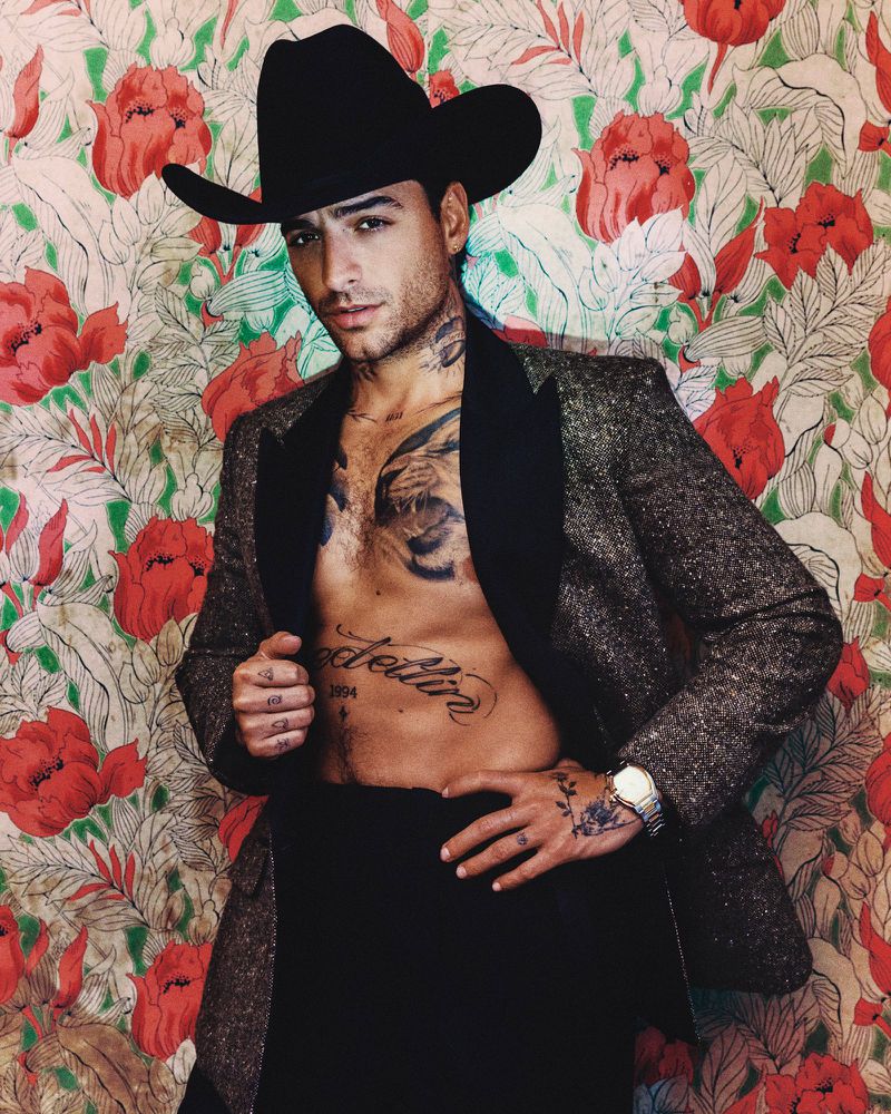 Maluma wearing a black cowboy hat and an open textured blazer showing chest tattoos against a floral wallpaper background.