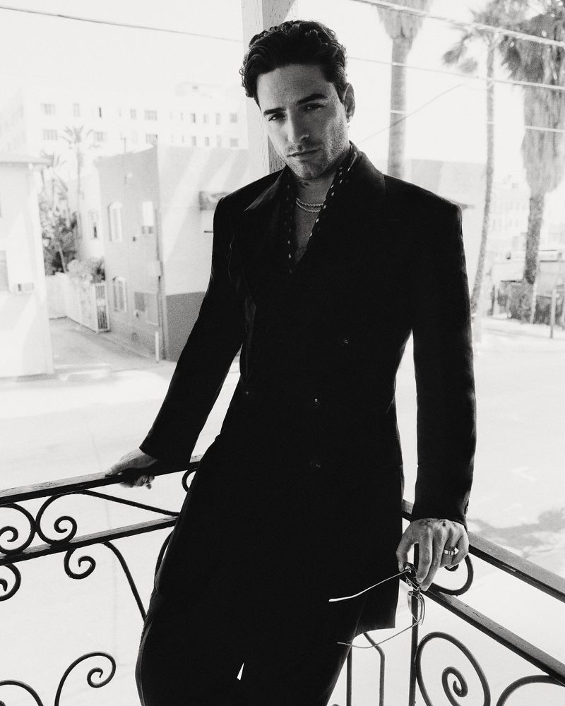 Man wearing a dark double-breasted coat leaning on a wrought-iron balcony railing in a monochrome portrait.