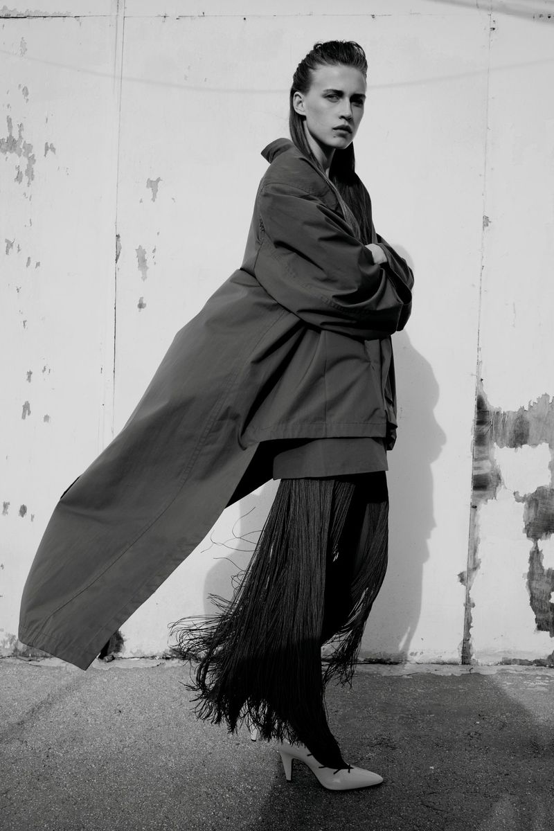 Model Liva Been poses in an oversized coat and long fringe trousers against a weathered wall in black and white.