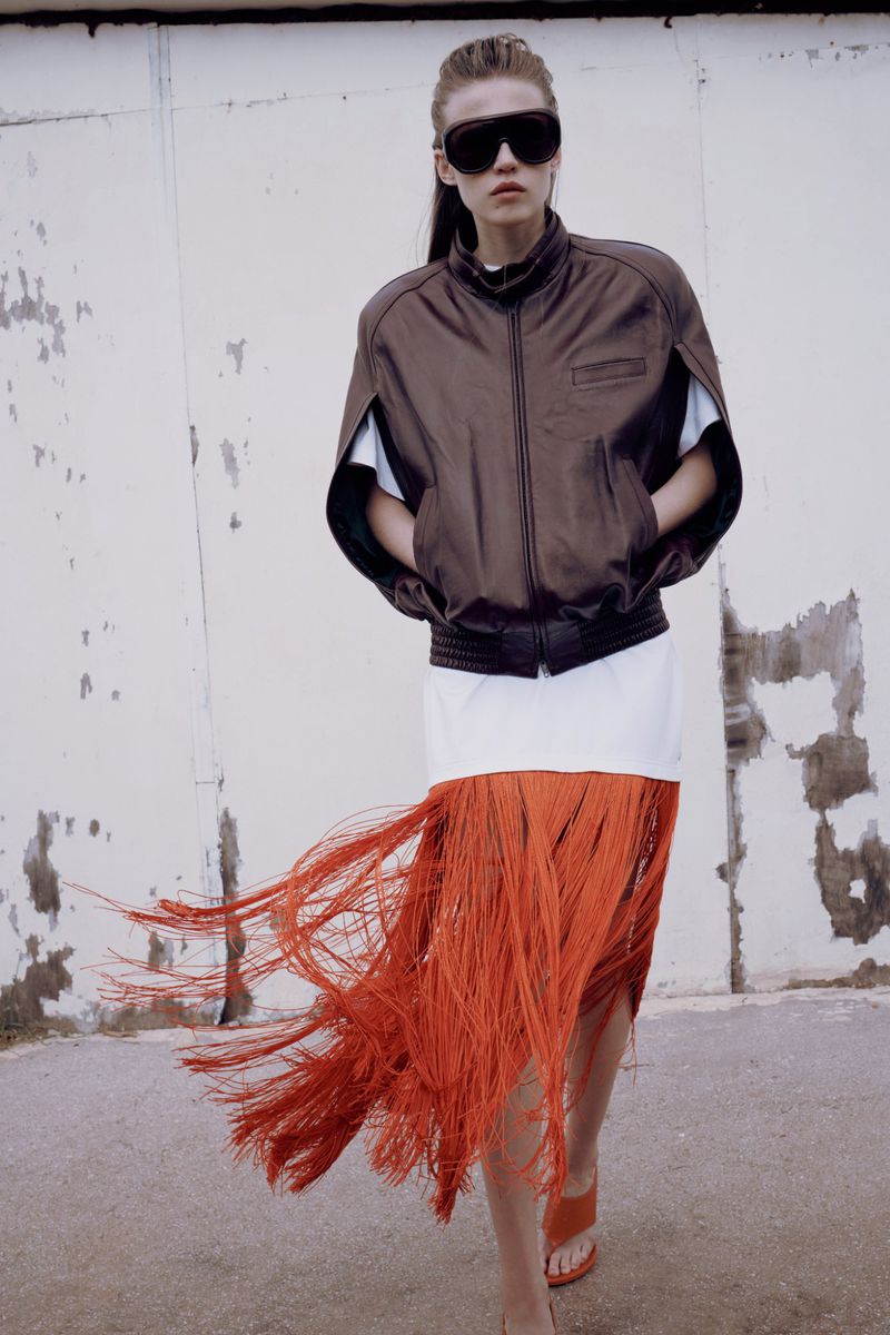 Model Liva Been in a brown jacket, large dark sunglasses, and a sweeping orange fringe skirt for Vogue Greece.
