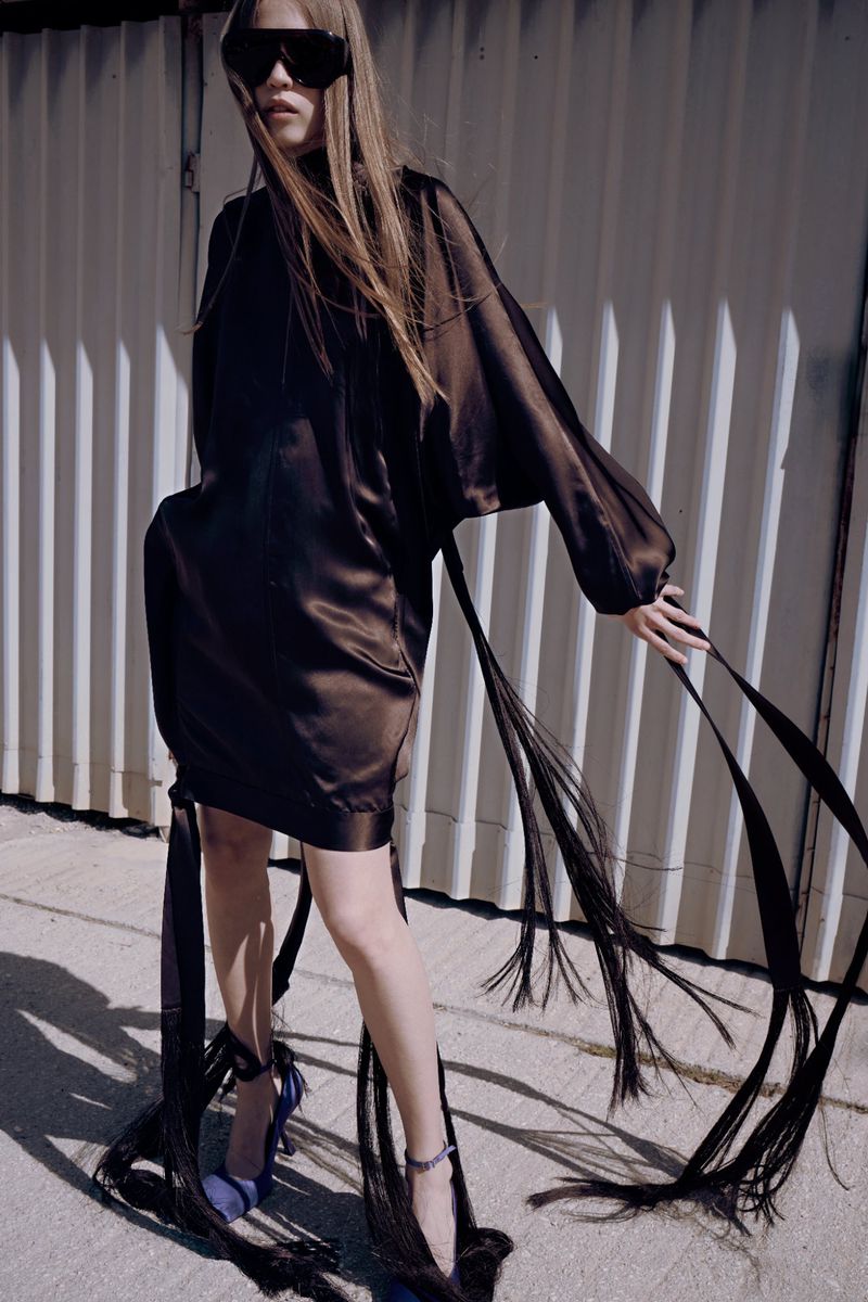 Model Liva Been wearing a dark brown satin dress with trailing fringe and black shield sunglasses against a corrugated wall.