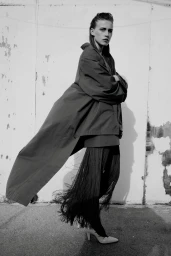 Model Liva Been poses in an oversized coat and long fringe trousers against a weathered wall in black and white.