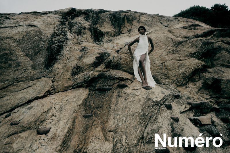 Model Larissa Joseph posing in a flowing white high-neck dress on a rugged rocky cliff face for Numéro Netherlands.
