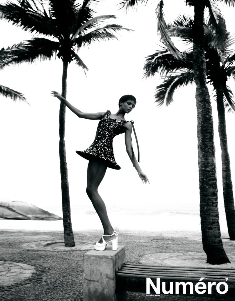 Model Larissa Joseph posing on a stone bench in a sequined mini dress and platform heels between palm trees on a beachfront promenade.