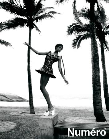 Model Larissa Joseph posing on a stone bench in a sequined mini dress and platform heels between palm trees on a beachfront promenade.