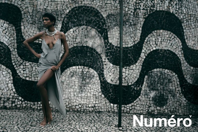 Model Larissa Joseph wearing a draped slip dress and layered necklace posing in front of a monochromatic wave mosaic wall.