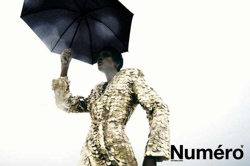 Model Larissa Joseph holding an open black umbrella while wearing a gold sequin coat against a bright white overcast sky.
