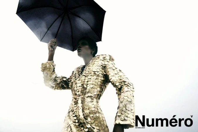 Model Larissa Joseph holding an open black umbrella while wearing a gold sequin coat against a bright white overcast sky.