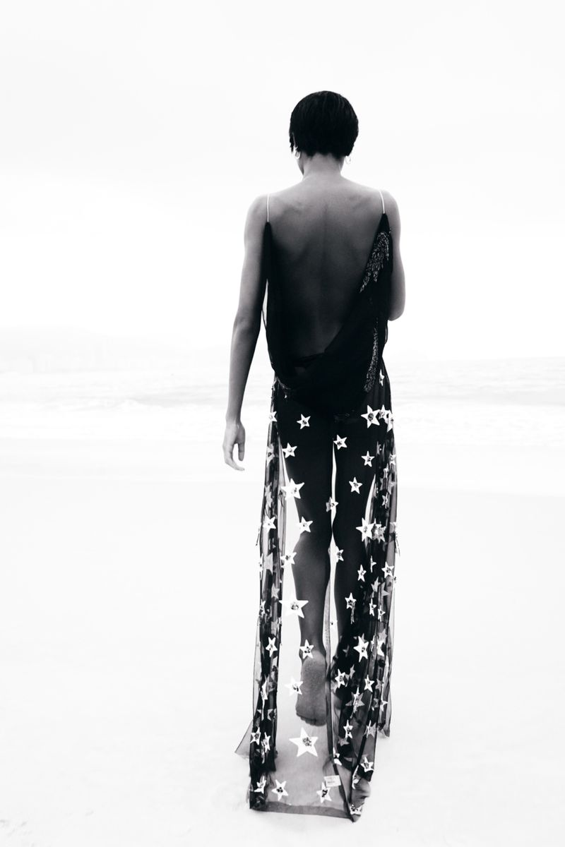 Model Larissa Joseph walking away on a sandy beach wearing a sheer backless black dress with star motifs.