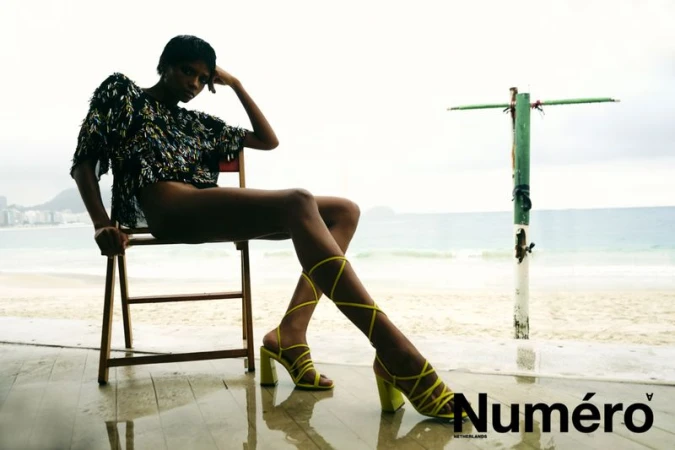 Model Larissa Joseph seated on a wooden chair in strappy lime heels and a dark textured top against a misty coastal beach backdrop.