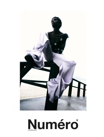 Model Larissa Joseph in wide-leg lavender silk trousers and a black embellished top posing on a dark railing near a beach.