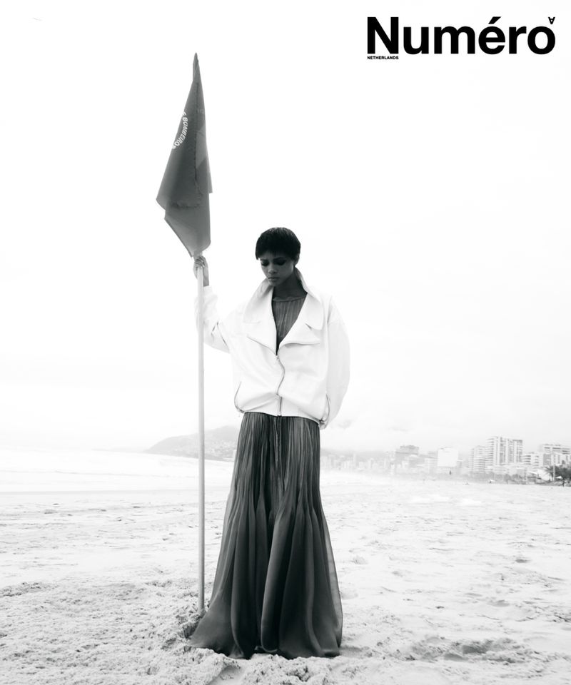 Model Larissa Joseph standing on a sandy beach holding a flag pole, wearing a white jacket and long pleated skirt in black and white.