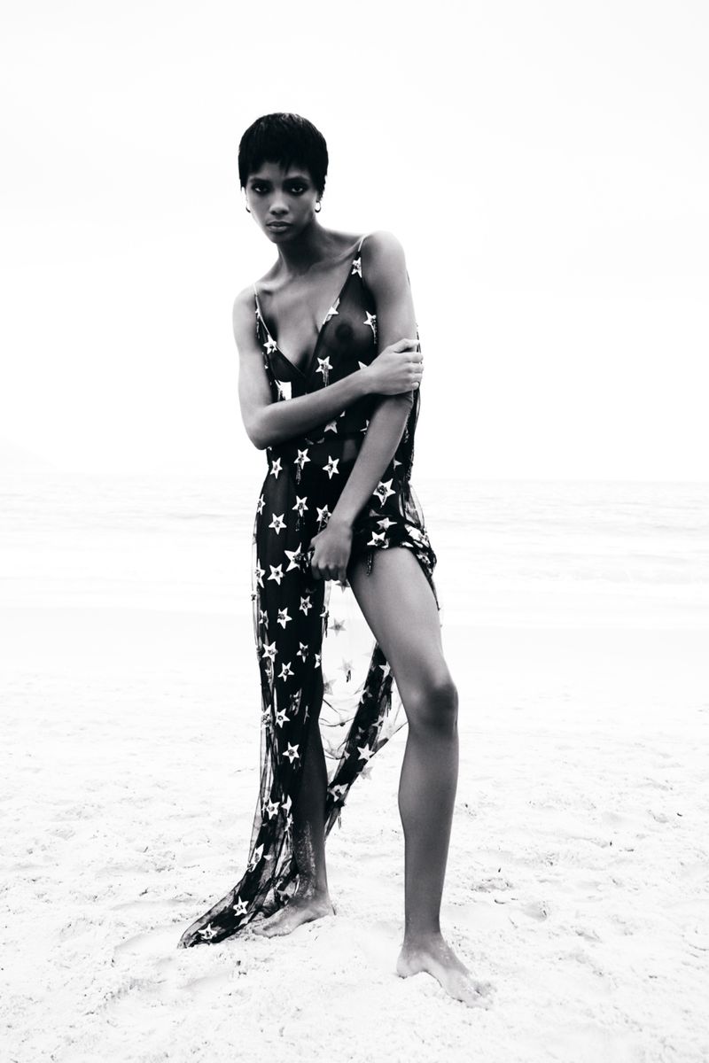 Model Larissa Joseph in a sheer star-patterned black dress standing barefoot on a bright sandy beach in black and white.
