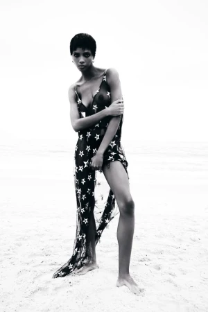 Model Larissa Joseph in a sheer star-patterned black dress standing barefoot on a bright sandy beach in black and white.
