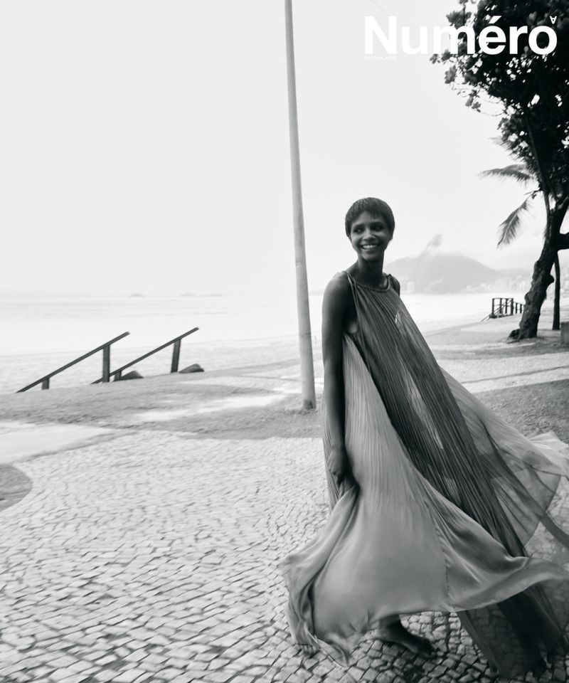 Black and white editorial photo of a smiling woman in a flowing pleated gown walking barefoot along a seaside cobblestone promenade.