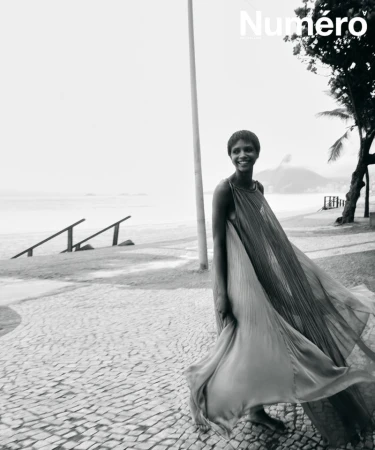 Black and white editorial photo of a smiling woman in a flowing pleated gown walking barefoot along a seaside cobblestone promenade.