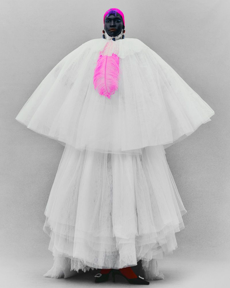 Person in a layered white tulle gown and cape with a pink feather embellishment, wearing a pink headwrap and black jewelry, studio shot.