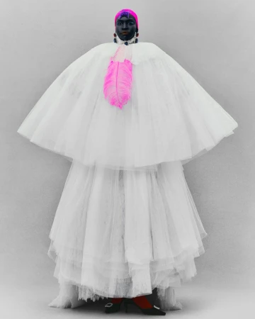 Person in a layered white tulle gown and cape with a pink feather embellishment, wearing a pink headwrap and black jewelry, studio shot.