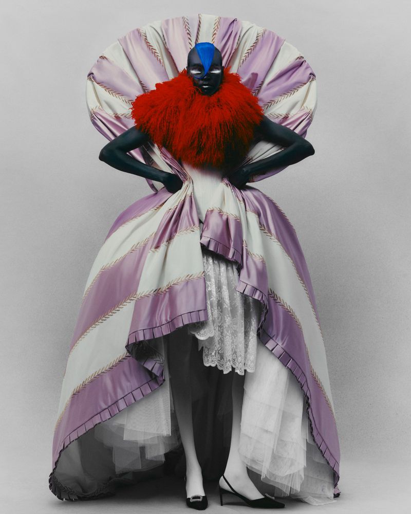 Striking fashion portrait of a person with blue hair and dark skin wearing a large striped gown with red feathered collar on white backdrop.