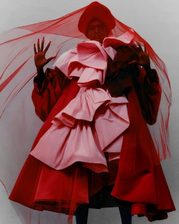A dark-skinned person is wearing an extravagant layered pink and red gown and matching head covering, set against a gradient grey backdrop.