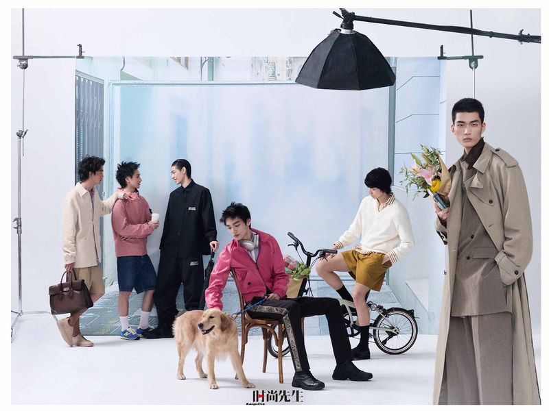 Several male models pose across an open studio set with a printed backdrop, a bicycle, studio lighting equipment, and a dog.