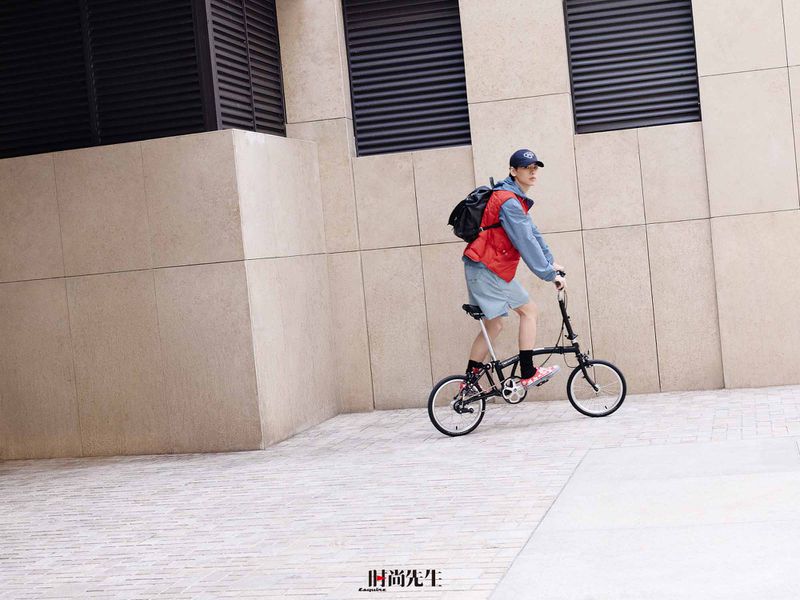 Man riding a black folding bicycle beside a modern beige building wearing a red puffer vest and blue cap.