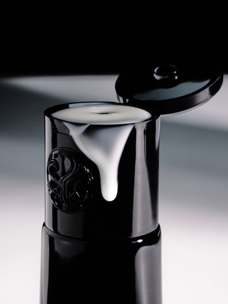 Glossy black cosmetic bottle with open flip-top cap showing thick white cream dripping down the front against a studio gradient.