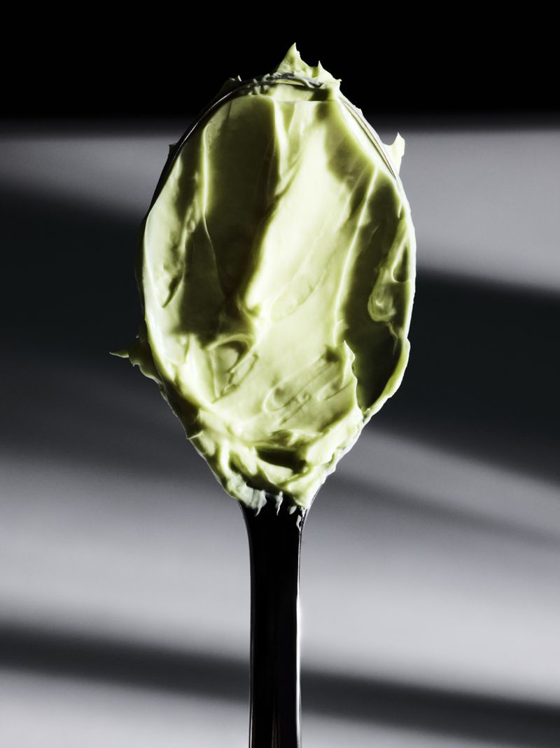 Close-up of pale green skincare cream textured on a black spoon against a striped grey and black background.