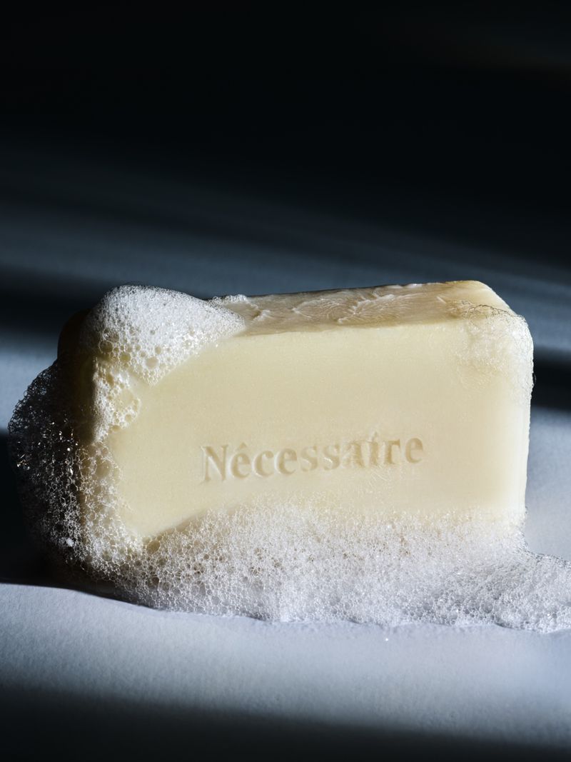 A cream-colored bar of Nécessaire soap covered in white suds and foaming lather under dramatic lighting.