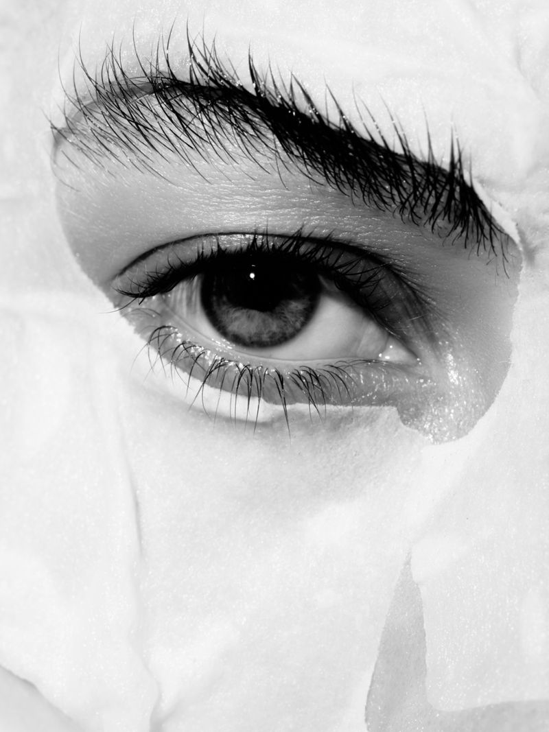 Black and white close-up of a person's eye and dark eyebrow looking through a jagged cutout in white torn paper.