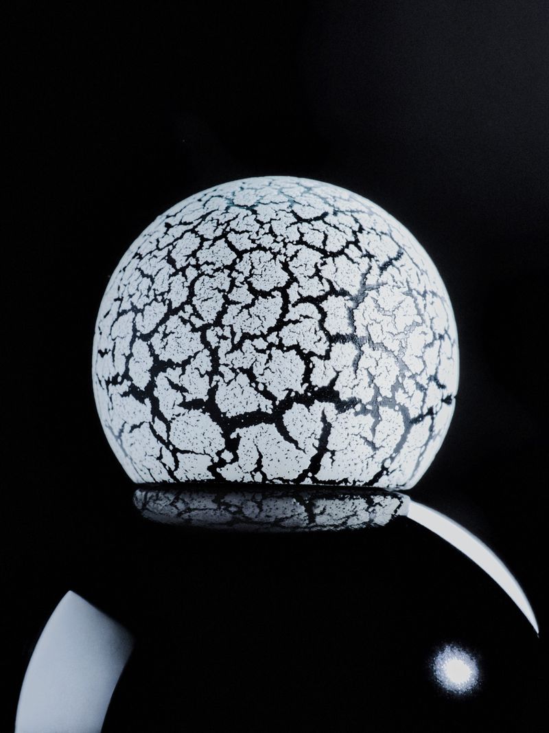 A white crackle-textured spherical dome resting on a glossy black reflective sphere against a solid dark background.