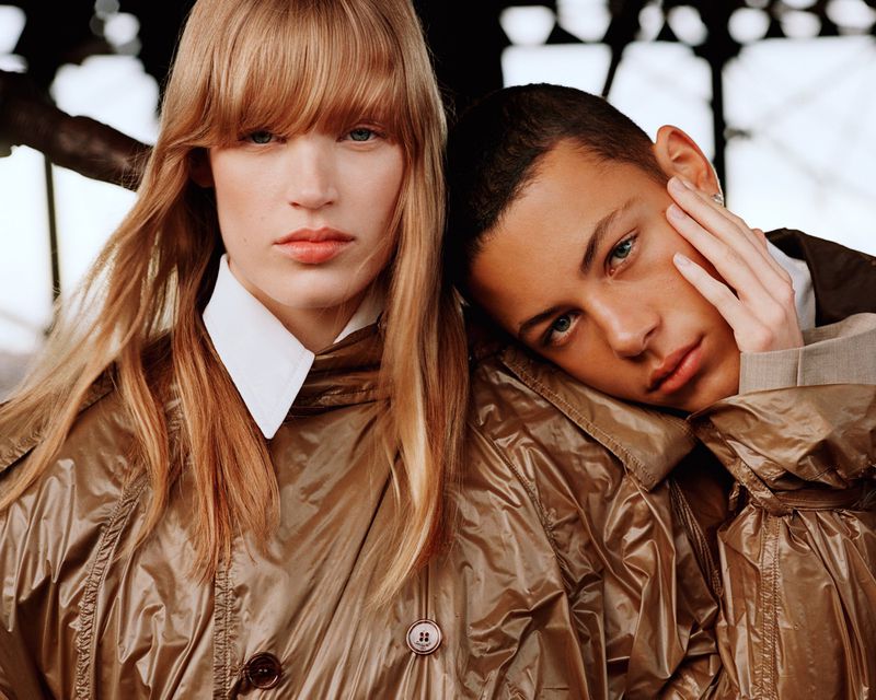 Close-up portrait of two models wearing shiny outerwear with a dark structural background.