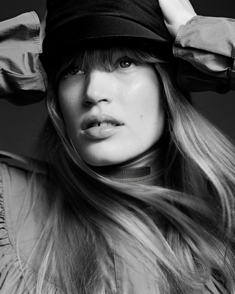 Black and white close-up portrait of a woman wearing a dark cap and high-neck top, with hands raised to her head.