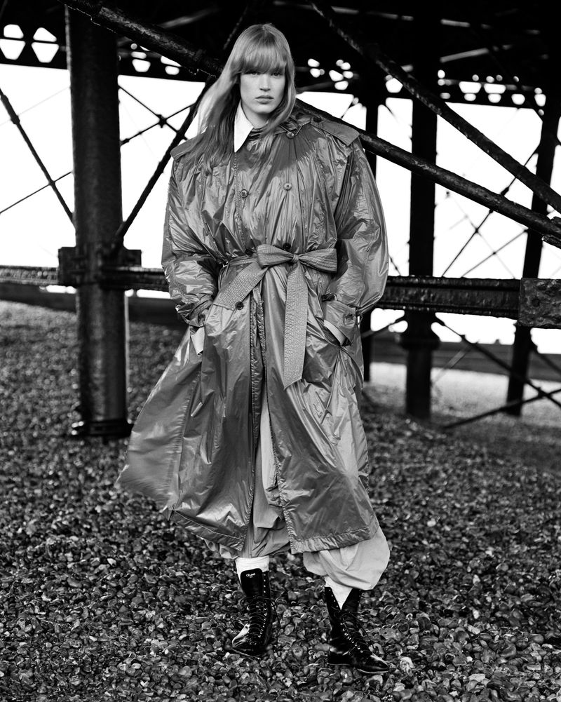 Jacqui Hooper wearing an oversized belted trench coat and boots while standing under an iron structure in a black and white shot.