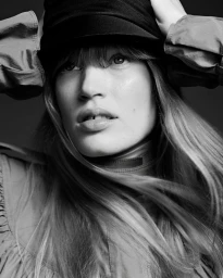 Black and white close-up portrait of a woman wearing a dark cap and high-neck top, with hands raised to her head.
