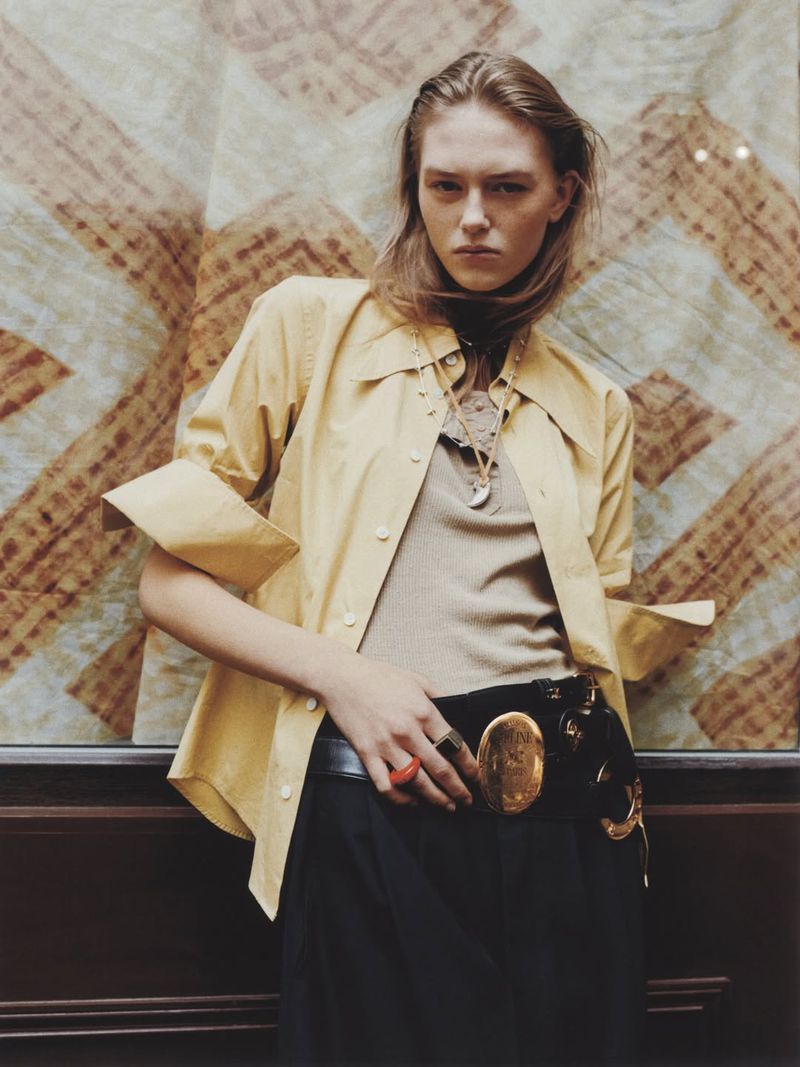 Model Agnes Wahlström in a pale yellow open shirt and a wide belt with an ornate gold buckle against a patterned textile backdrop.