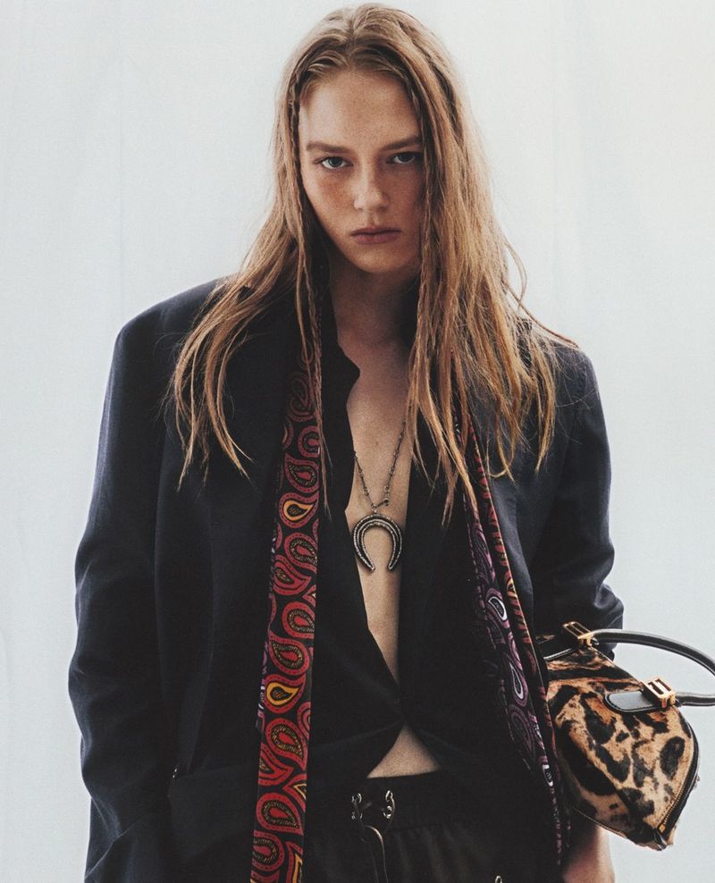 Model Agnes Wahlström wearing an open black blazer, paisley scarf, and crescent pendant holding a leopard print handbag against a pale backdrop.