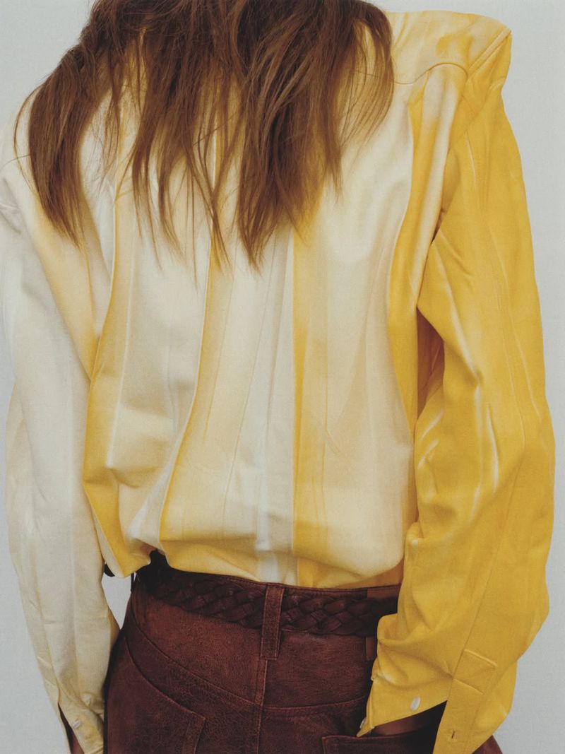 Rear view of a model wearing a structured yellow and white gradient shirt with brown pants against a plain white studio backdrop.